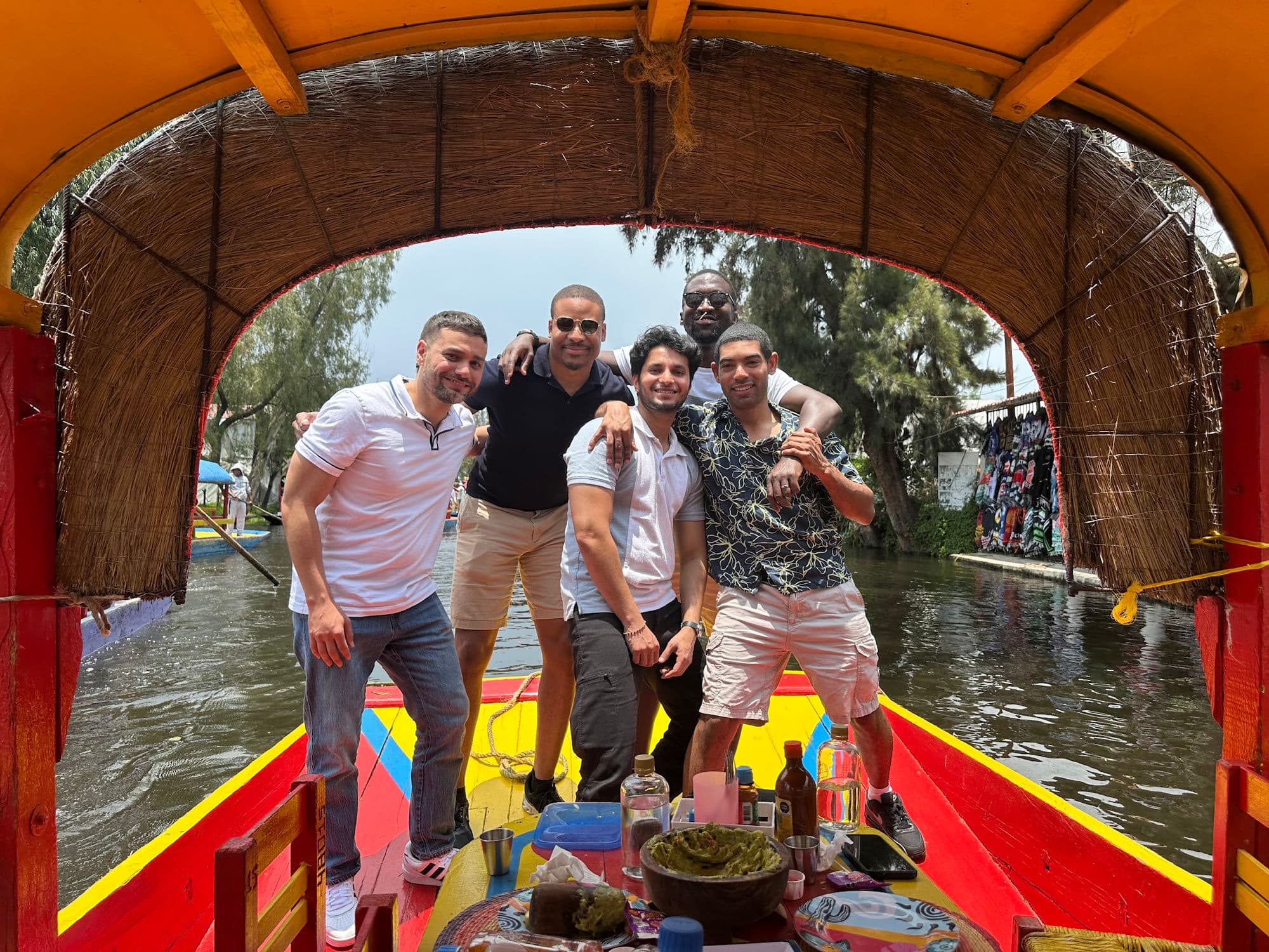 Felipe and four friends posing together on a colorful boat in Xochimilco, Mexico City, with guacamole and drinks on the table in front of them.